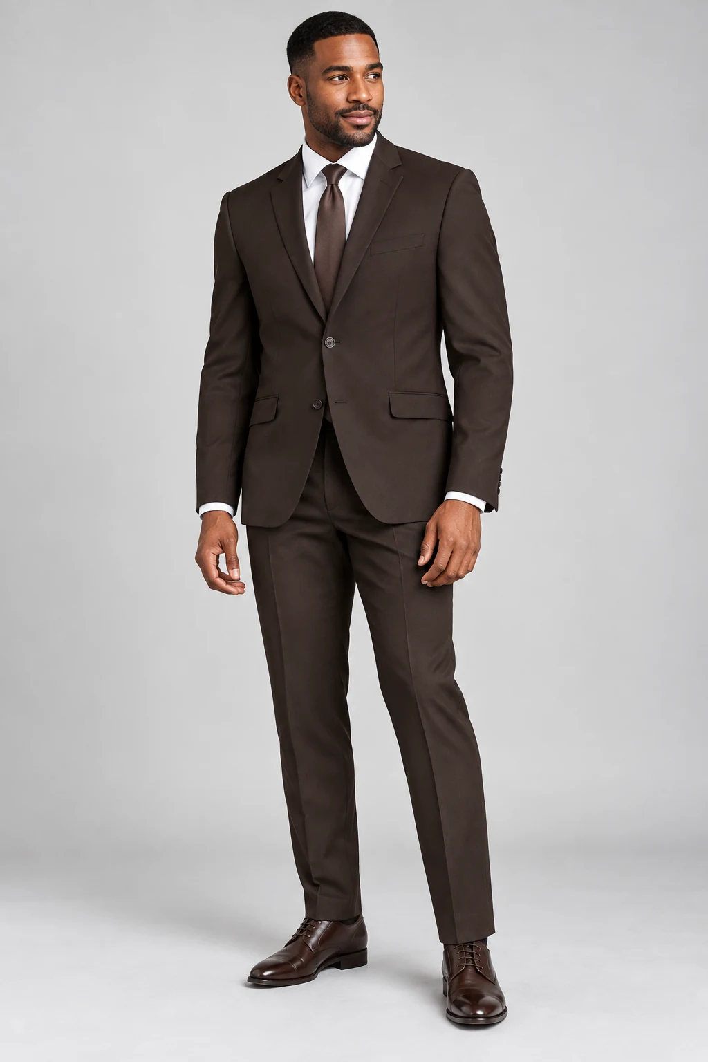 Model wearing a dark brown two-button suit, white shirt, brown tie and brown leather shoes on a plain gray studio background.